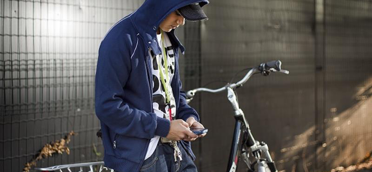 Jongen met mobieltje in de hand leunt tegen zijn fiets en heeft en capuchon op
