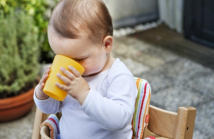 Kindje zit in de kinderstoel in de tuin en drinkt uit een plastic beker