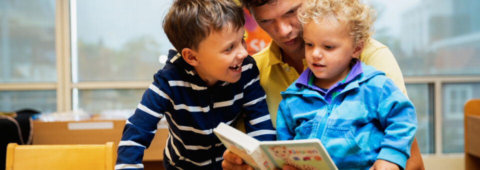 Vader is met een zoon op schoot en een jongetje ernaast aan het voorlezen in de bibliotheek