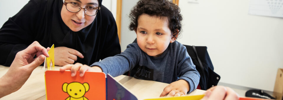 Jongetje kijkt naar een boekje en houdt dit vast terwijl zijn moeder meekijkt tijdens het ontwikkelingsonderzoek door de jeugdverpleegkundige van het CJG