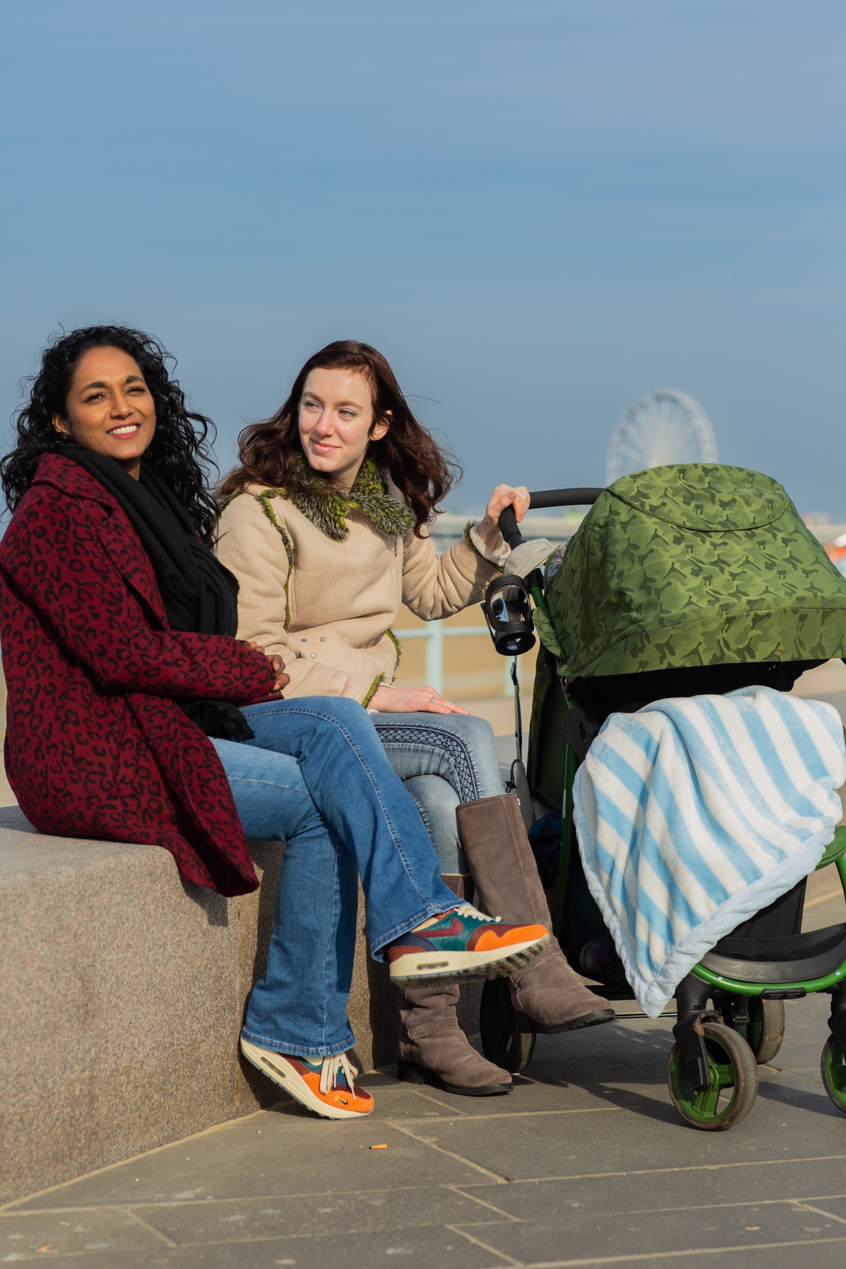 Twee MIM moeders zitten op de boulevard van Scheveningen met naast elkaar in de zon en er staat een kinderwagen naast